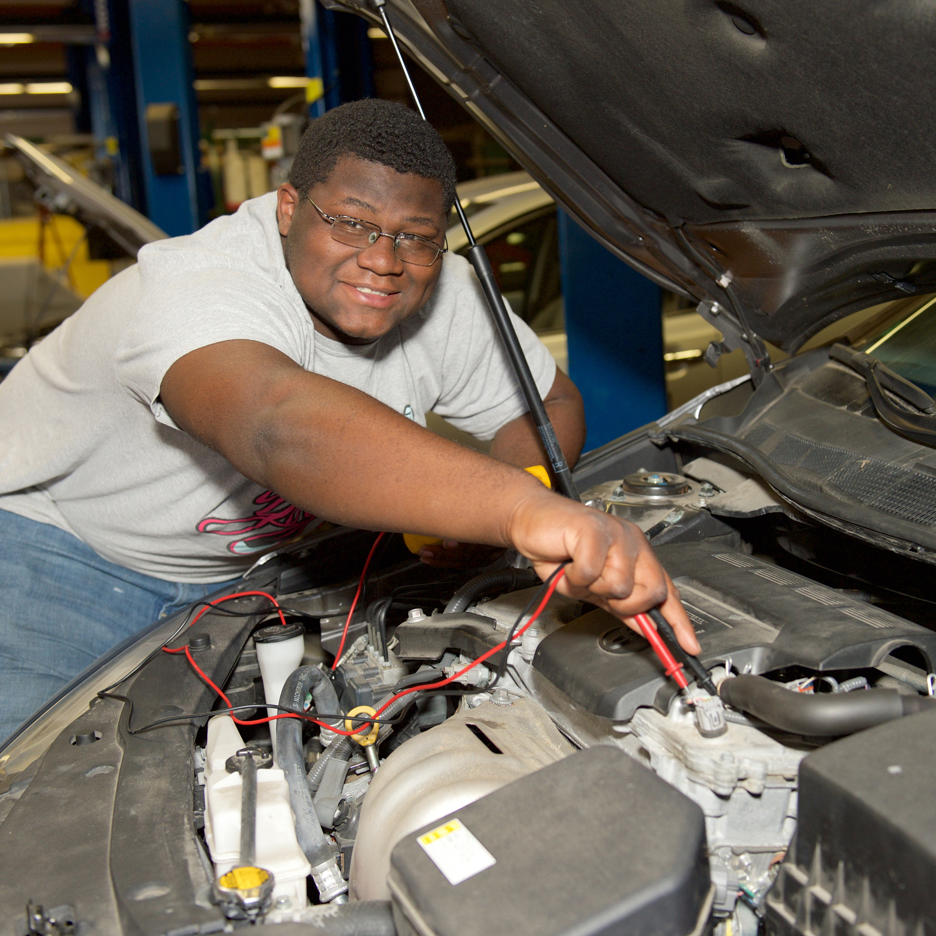 Auto student working on a car engine holding the red and black cords of an electrical meter to take a reading.