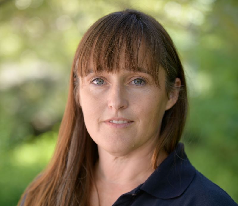 Megan Rhodes in blue shirt in front of blurred green background of trees and shrubs