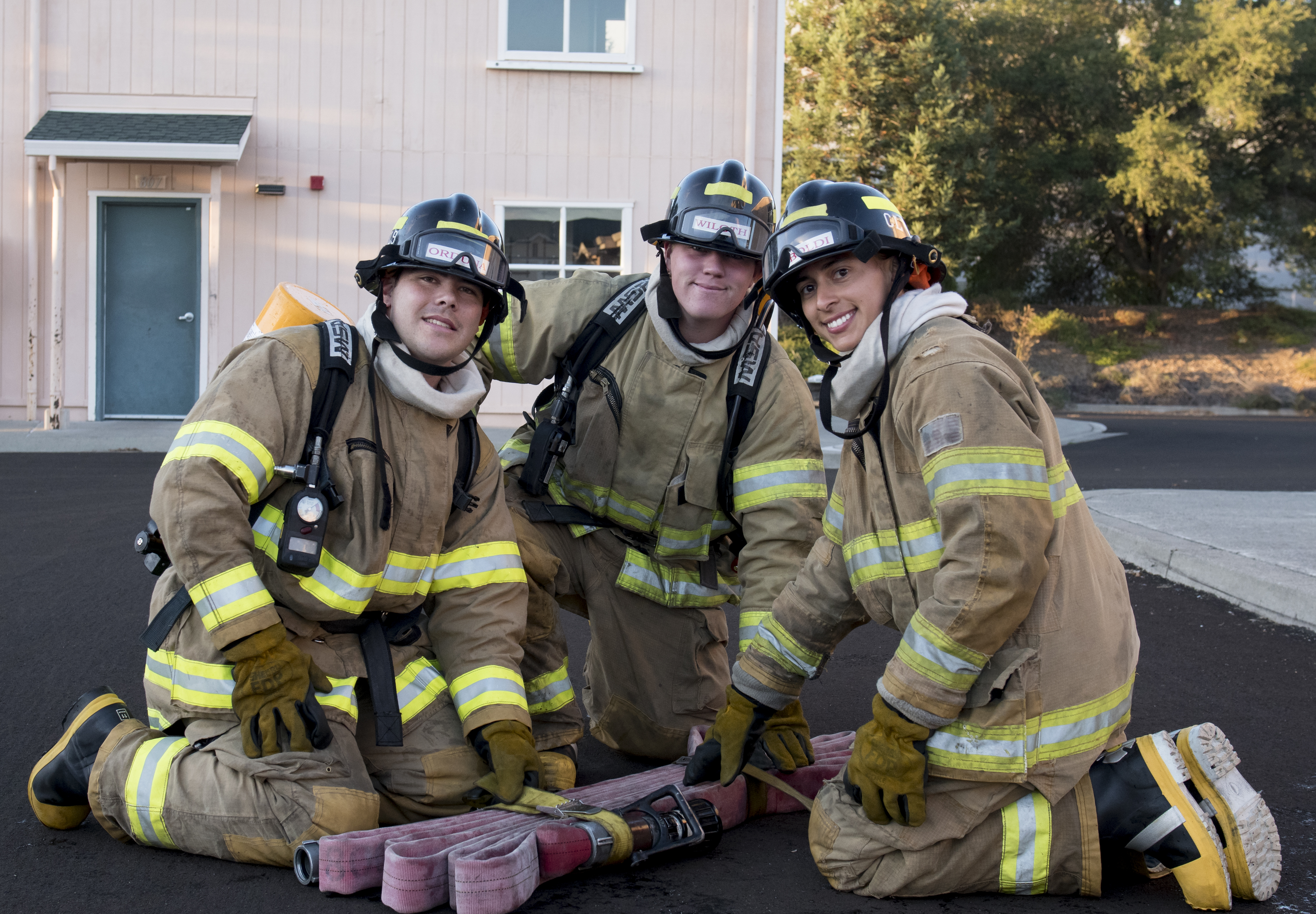 Three firefighter trainees in a group wearing their firefighting gear kneeling with a firehose between them